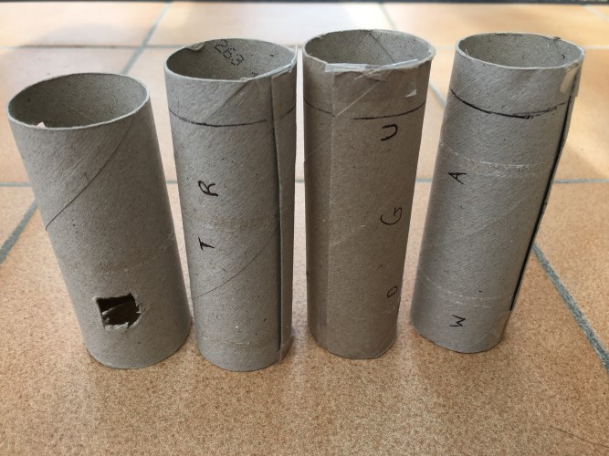 An unassembled cryptex made from toilet tubes with letters written randomly across them, and one tube with a hole cut into it so that when it is slid onto the other tubes letters are revealed in the hole